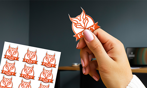 Hand holding “FOXY” sticker with fox head design — identical sticker sheet and desk setup visible in background.
