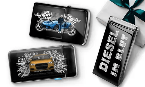 Two sleek black lighters featuring car themes, one with a blue car and checkered flags, another with a red car and gears. A third lighter reads 'DIESEL IM BLUT'. Gift-wrapped in the background.