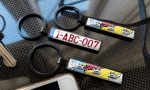 Llaveros de matrícula en estilo cómic, exhibidos sobre una superficie con textura de carbono y diseños de coches coloridos